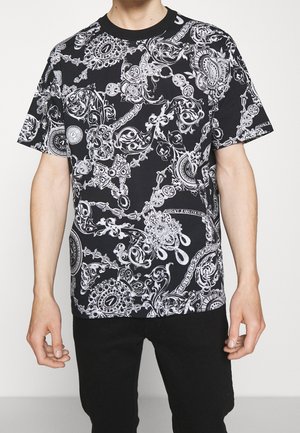 T-shirt noir à manches courtes avec des motifs baroques blancs complexes, des chaînes et des motifs floraux, porté par une personne avec un pantalon noir.