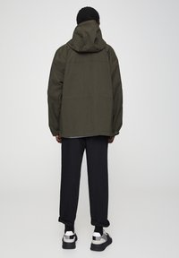 Olijfgroene, gevoerde jas met een losse pasvorm en licht gestructureerde stof, gecombineerd met zwarte broek en sportieve zwart-witte sneakers.