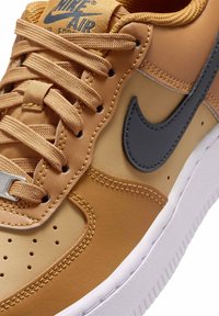 Buty Nike Air Force 1 w kolorze tan z szarymi akcentami, charakteryzujące się gładką teksturą, perforowanym czubkiem oraz plecionymi sznurówkami. Biała podeszwa.