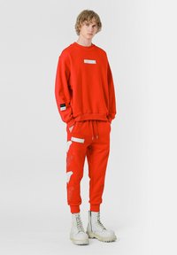 Sudadera roja y pantalones jogger a juego con gráficos de plumas blancas y detalles plateados. El modelo lleva botas blancas gruesas. Estilo de ropa deportiva casual.