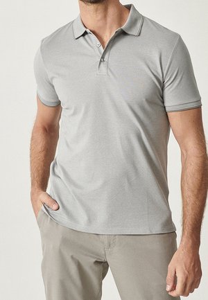 Man draagt lichtgrijs poloshirt met korte mouwen en knoopkraag en beige broek, één hand in de zak tegen een effen achtergrond.