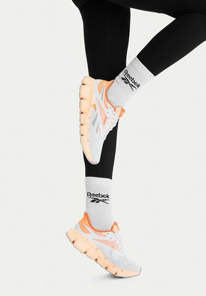 Nogi w czarnych legginsach, na stopach białe i brzoskwiniowe buty do biegania Reebok oraz białe skarpetki z logo Reebok, jedna noga zgięta i uniesiona.