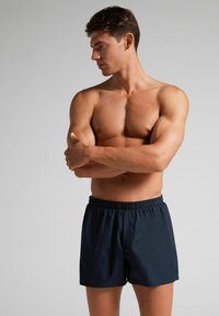 Námořnické plavky s elastickým pasem a hladkým povrchem. Model prezentuje boční profil, paže překřížené.