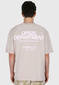 Világosszürke, nagyméretű póló, fehér felirattal a hátsó részén: "DPSZE DEPARTMENT," "MADE FOR ELITE HUSTLERS©," és "DROPSIZE WORLDWIDE."