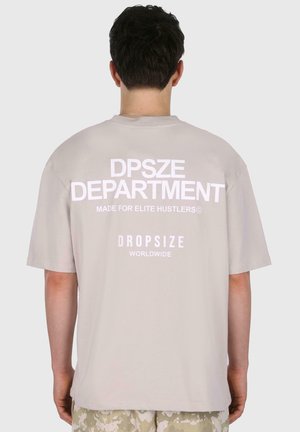 Lys grå oversized t-skjorte med hvit tekst på baksiden: "DPSZE DEPARTMENT," "MADE FOR ELITE HUSTLERS©," og "DROPSIZE WORLDWIDE."