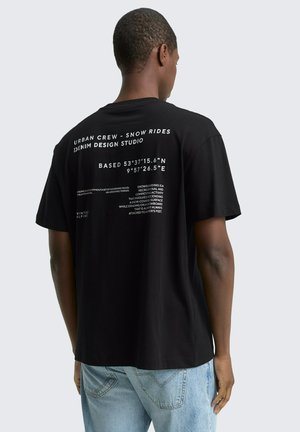 Mann trägt schwarzes T-Shirt mit weißem Text auf dem Rücken, der Koordinaten und snowboardbezogene Sprüche zeigt, kombiniert mit hellblauen Jeans.