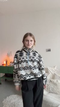Fleece trui in zwart en wit met geometrische patronen, hoge kraag en halve rits. Draagt losse zwarte broek en staat in een gezellige interieur.