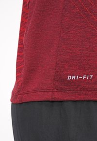 Camiseta deportiva Dri-FIT roja con una superficie texturizada, que presenta un dobladillo curvado y costuras visibles. Tela negra debajo de la camiseta.