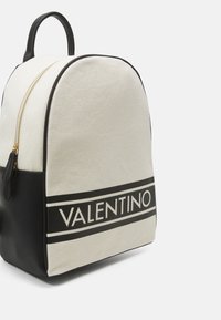Mochila de lona con forma redondeada. Color crema con una sección inferior negra y una banda horizontal negra resaltante que muestra "VALENTINO". Cierre dorado.