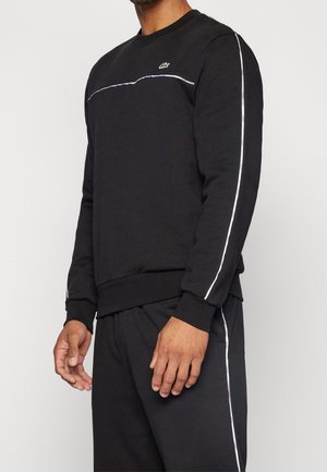 Muž v černé mikině a kalhotách s bílými reflexními pruhy na rukávech a nohavicích kalhot, logo Lacoste na hrudi mikiny.