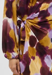 Vestito colorato con una combinazione di sfumature viola, giallo e bianco, caratterizzato da motivi astratti, maniche lunghe e un dettaglio annodato in vita.