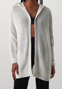 Bílý pletený cardigan s volným, otevřeným designem, který má kapuci, žebrované manžety a texturovaný vzor po celé ploše.