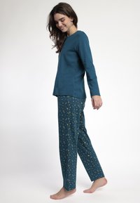 Mujer sonriendo, caminando descalza, vistiendo una blusa de manga larga color azul verdoso y pantalones a juego de color azul verdoso con patrones celestiales dorados.