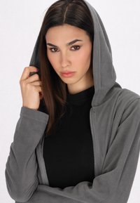 Felpa grigia con zip e cappuccio, caratterizzata da una superficie liscia e una cerniera frontale. Indossata sopra una maglietta nera, presenta un design semplice.