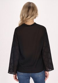 Schwarze Bluse mit langen, transparenten Spitzeärmeln in einem diamantförmigen Muster. Glatte Textur, runder Ausschnitt und lockere Passform.