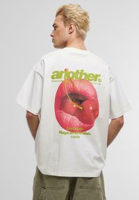 White cotton t-shirt with a graphic print of red lips and cherries, featuring green text that reads "another" and "Un Club de Playa en el Paraíso."