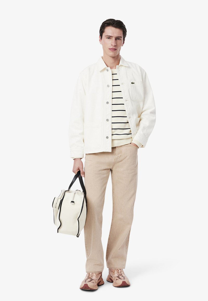 Junger Mann trägt eine weiße Jacke über einem gestreiften Pullover, beige Hose, pinke Sneaker und hält eine weiße und schwarze Tragetasche.