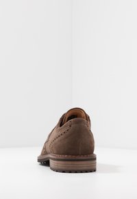 Chaussure habillée pour homme en daim marron avec détails brogue et talon empilé, vue de dos sur une surface blanche avec un fond gris clair.