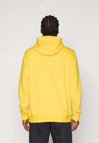 Sweatshirt amarelo com um capuz espaçoso, punhos e bainha canelados. Textura suave, ajuste descontraído, sem logótipos ou padrões visíveis. Calças escuras.