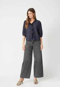 Blusa blu navy con collo arricciato e maniche a palloncino a tre quarti, abbinata a jeans grigi a vita alta e pantaloni larghi, e sandali verde chiaro.