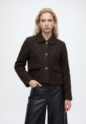 Femme aux cheveux blonds ondulés portant une veste marron foncé texturée avec des boutons dorés et un pantalon en cuir noir, debout devant un fond uni.