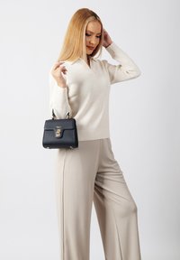 Schwarze handgefertigte Handtasche mit goldenen Beschlägen, in einer Hand gehalten. Das Modell trägt einen hellbeigen Pullover und cremefarbene Hosen. Einfaches Design, minimale Details.
