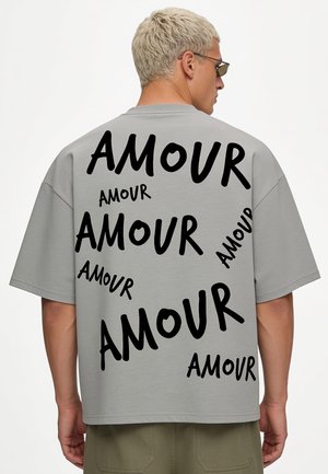 Persona che indossa una maglietta grigia oversize con la parola "AMOUR" stampata più volte in varie dimensioni e direzioni sulla schiena.