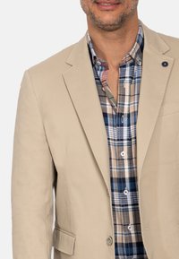 Blazer beige con dos botones, llevado sobre una camisa de cuadros en azul, marrón y blanco. Tela suave con un ligero brillo, ajuste entallado.