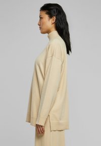 Beige-colour turtleneck-paita, jossa on pitkät, löysät hihat, sivuaukot ja ribbaatut hihansuut. Pehmeä rakenne ja rento istuvuus keskipitkässä designissa.