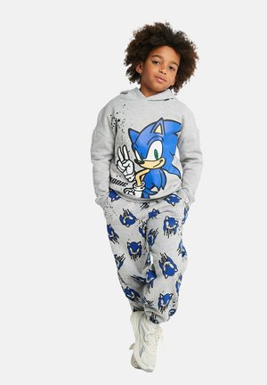 Child with curly hair wearing light gray hoodie and pants featuring blue Sonic the Hedgehog graphics, paired with white sneakers, standing relaxed.
