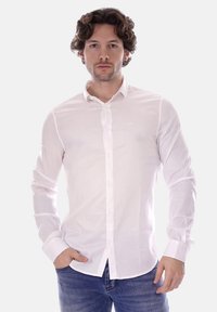 Camicia bianca a maniche lunghe con bottoni, slim fit, realizzata in tessuto morbido. Presenta un colletto e un logo discreto sul petto. Indossata con jeans blu.