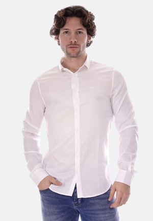 Camicia bianca a maniche lunghe con bottoni, slim fit, realizzata in tessuto morbido. Presenta un colletto e un logo discreto sul petto. Indossata con jeans blu.