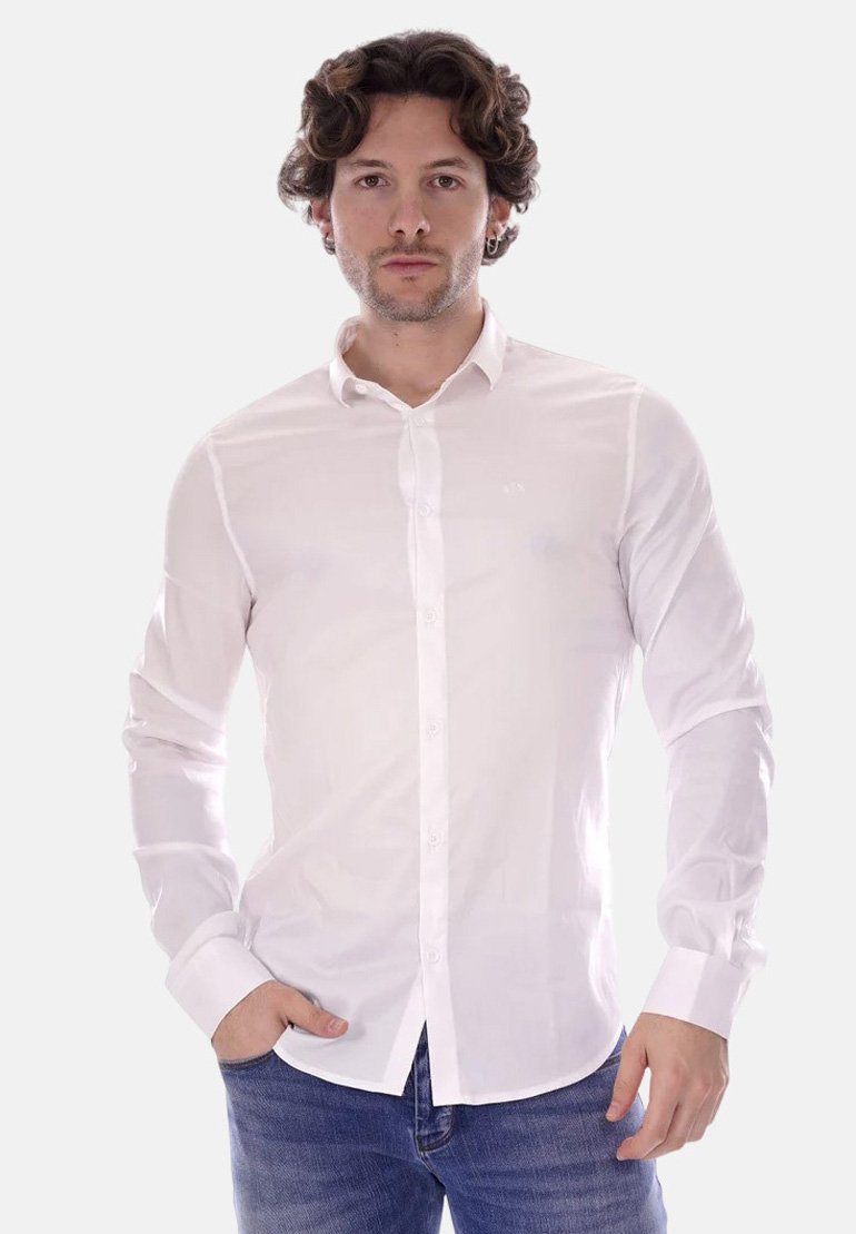 Camicia bianca a maniche lunghe con bottoni, slim fit, realizzata in tessuto morbido. Presenta un colletto e un logo discreto sul petto. Indossata con jeans blu.