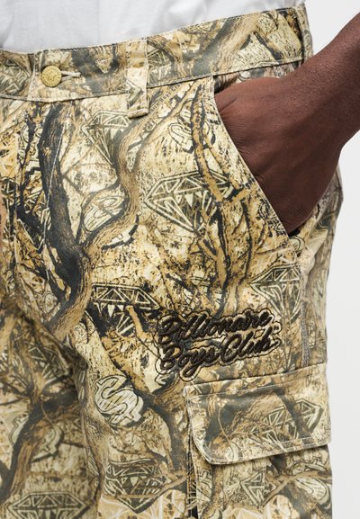 Μπεζ cargo σορτς με καμουφλάζ σχέδιο που περιλαμβάνει δέντρα και διαμάντια. Διαθέτει πλαϊνή τσέπη και ανάγλυφο λογότυπο με μαύρη ραφή.