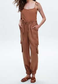 Haut marron sans manches associé à un pantalon cargo ample, doté d'une ceinture élastique et de poches latérales. Le modèle porte des sandales marron à enfiler.