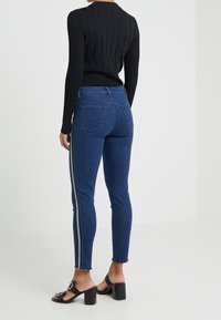 Skinny jeans in dark blue denim with a contrasting silver side stripe. Features five pockets and a fitted silhouette. Worn with a black ribbed top.