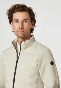Beige zip-up jacka med quiltad front och strukturerade, enfärgade ärmar. Har en svart cirkulär logotyp på vänstra ärmen. Slät yta.