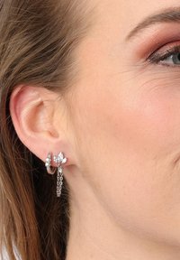 Zilveren oorbellen met een bloemmotief, grote heldere edelstenen en een kettingaccent. Toont een close-up van de oorbel aan het oor van een model.