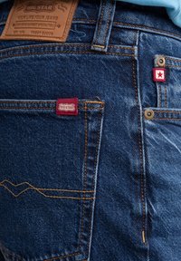 Dunkelblaue Jeans aus Denim mit orangefarbenen Nähten, ausgestattet mit einem Lederpatch, einem roten Stoffetikett und einem Metallnieten an der hinteren Tasche.