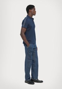 Hombre de perfil con una polo azul marino de mangas estampadas, jeans azules y zapatos negros, contra un fondo liso.