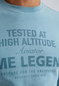 Helles blaues Baumwoll-T-Shirt mit dunkelblauen Schriftzug: "GETESTET IN HOHER HÖHE", mit verschiedenen Schriftgrößen und -stilen. Rundhalsausschnitt.