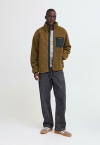 Young man standing wearing brown fleece jacket with black chest pocket, gray t-shirt, loose black jeans, and tan sneakers with white stripes.