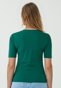Vrouw met schouderlang haar, gekleed in een groene T-shirt met korte mouwen, gezien van achteren, gecombineerd met licht denim shorts tegen een neutrale achtergrond.