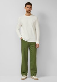 Man staat met één hand in de zak, gekleed in een wit overhemd met lange mouwen, olijfgroene broek en witte sneakers, tegen een grijze achtergrond.