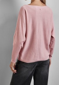 Jasnoróżowy sweter z miękkiego materiału o szerokim dekolcie i długich rękawach, z małym detalem na plecach. Połączony z ciemnoszarymi spodniami.