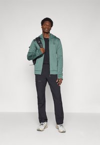 Grüne Kapuzenjacke mit Reißverschluss und glatter Textur. Kombiniert mit schwarzen Hosen und Schuhen, dazu ein Rucksack mit mehrfarbigen Riemen.