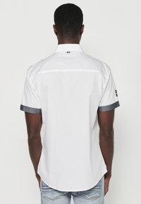 Camisa blanca de manga corta con diseño de cuello, que presenta puños azul oscuro y un discreto detalle de costura en la parte posterior.