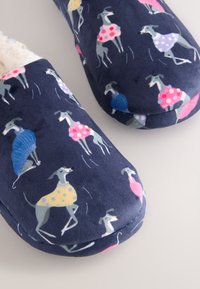 Marineblaue Hausschuhe mit einem Muster aus grauen Hunden, die bunte Pullover und Kleider in Pink, Gelb, Blau und Lila tragen.