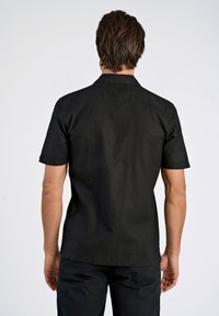 Chemise noire à manches courtes en tissu léger. Dotée d'un col, de manches retroussées et d'une coupe décontractée. Pas de motifs visibles.