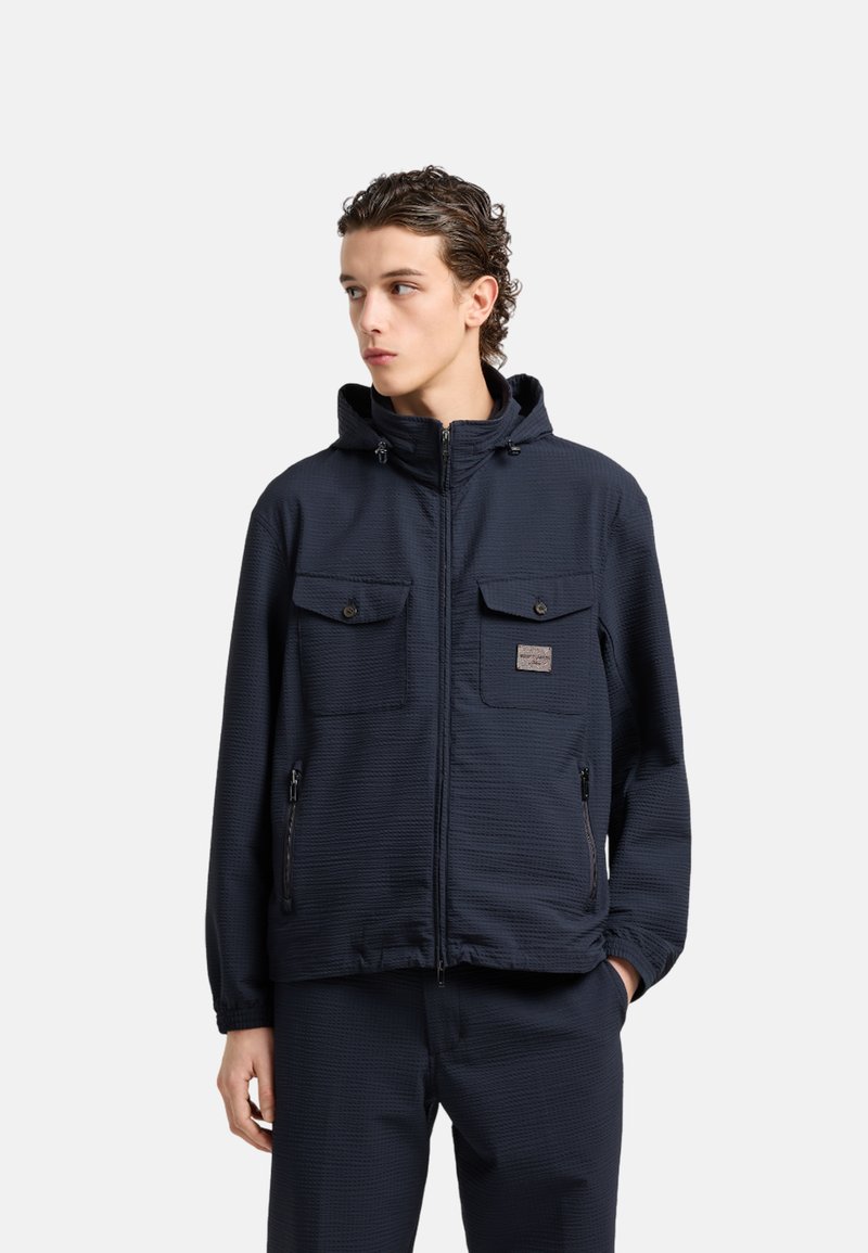 Giacca zip-up blu navy con tessuto testurizzato, dotata di cappuccio, due tasche sul petto con pattina e tasche laterali con zip.